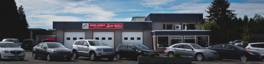 Row of pre-owned cars and suvs in front of a 3-bay garage. Auto sales office on right side.